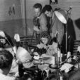 Two men in suits record the actions of women doing sewing work in a factory