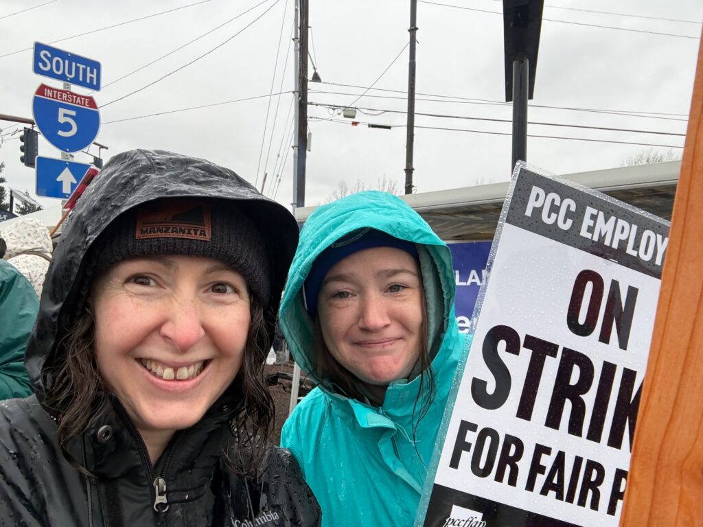 Two wet women in raincoats on a rainy day. One is holding a sign that says PCC Employees on strike for fair pay.
