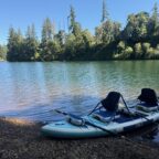 Kayak and two paddles next to a lake with green trees in a background and a blue sky